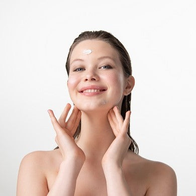 Brünette, lächelnde Frau fasst sich mit zwei Händen an den Hals und trägt Kleckse der Greek Yoghurt Gesichtspflege im Gesicht. 